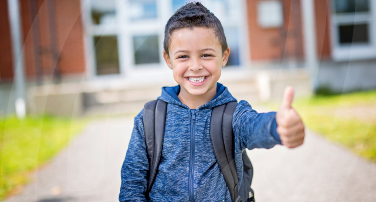Young child having thumbs up in front of school